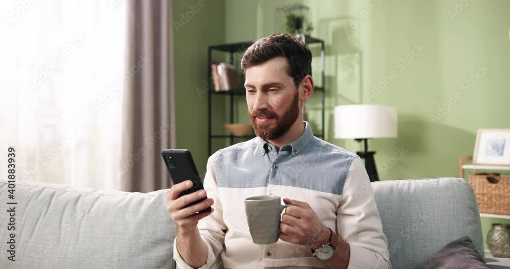Close up portrait of happy handsome Caucasian bearded man sitting on sofa in cozy home, smiling typing and texting on smartphone gadget while sipping coffee or tea in room. Leisure concept Close up portrait of happy handsome Caucasian bearded man sitting on sofa in cozy home, smiling typing and texting on smartphone gadget while sipping coffee or tea in room. Leisure concept