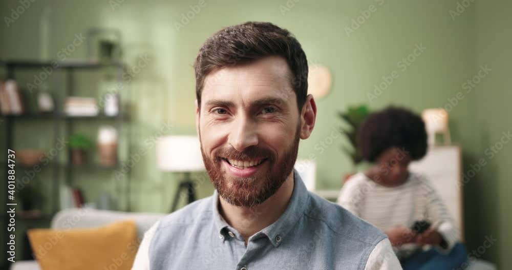 Close up portrait of joyful young bearded handsome young guy looking at camera and smiling at home in cozy room while African American woman sits behind. Leisure concept. positive emotions Close up portrait of joyful young bearded handsome young guy looking at camera and smiling at home in cozy room while African American woman sits behind. Leisure concept. positive emotions