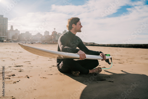 Surfer sitting on sandy beach with surfboard.