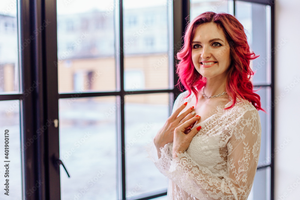 Attractive woman with pink hair in white light dress posing on near large window. Lady in gentle lace peignoir Attractive woman with pink hair in white light dress posing on near large window. Lady in gentle lace peignoir
