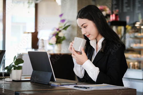 Side view of attractive businesswoman drinking coffee and working with computer tablet while sitting at in modern cafe.