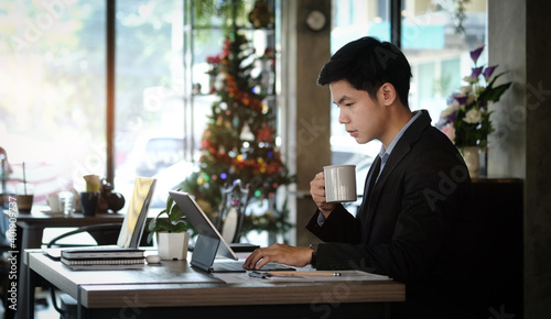 Businessman in suit working on tablet computer and writing in formation on document while sitting at modern coffee shop.