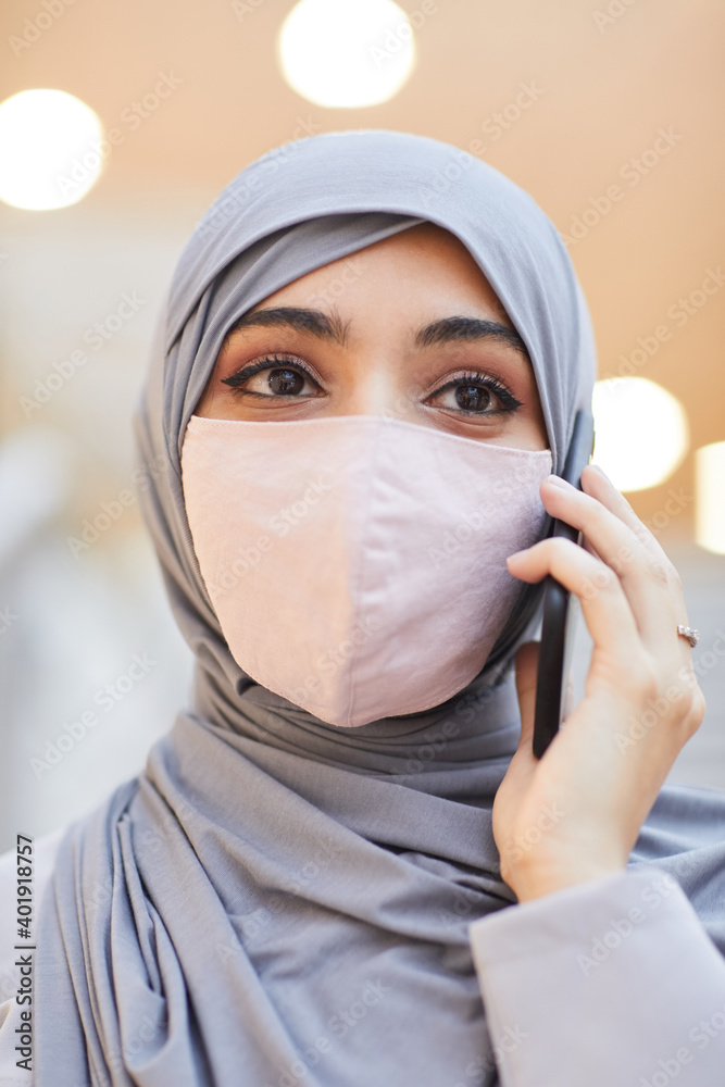 Vertical close up portrait of modern Middle-Eastern woman wearing mask and speaking by smartphone while standing in shopping mall