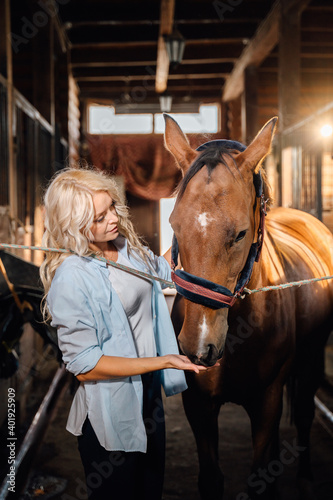 A pretty blonde dressed in a shirt stands in the stable next to a horse.