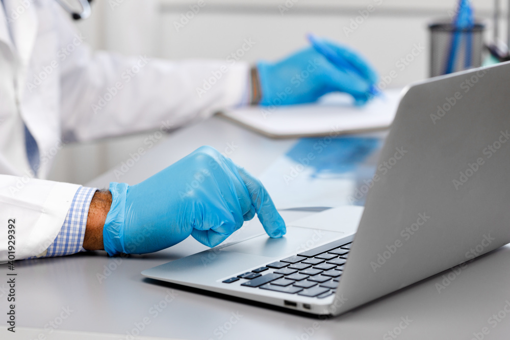 Doctor in medical gown and gloves typing on laptop and making notes at working table