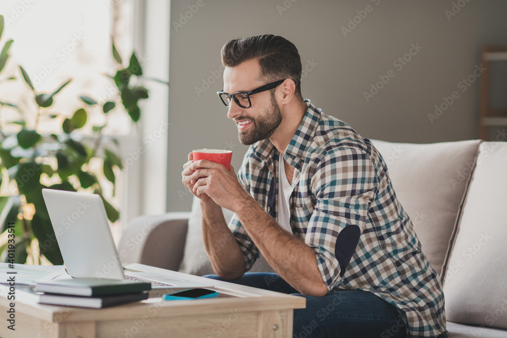 Profile side photo of young handsome man happy positive smile enjoy cup pf coffee break pause look laptop sit couch home Profile side photo of young handsome man happy positive smile enjoy cup pf coffee break pause look laptop sit couch home