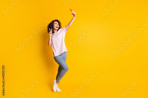 Full length body size photo of female millennial taking selfie showing v-sign isolated on vivid yellow color background with copyspace
