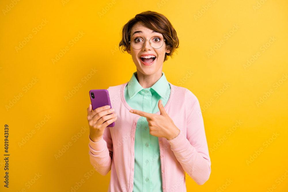 Photo portrait of excited girls wearing glasses pointing with finger on smartphone isolated on bright yellow color background Photo portrait of excited girls wearing glasses pointing with finger on smartphone isolated on bright yellow color background