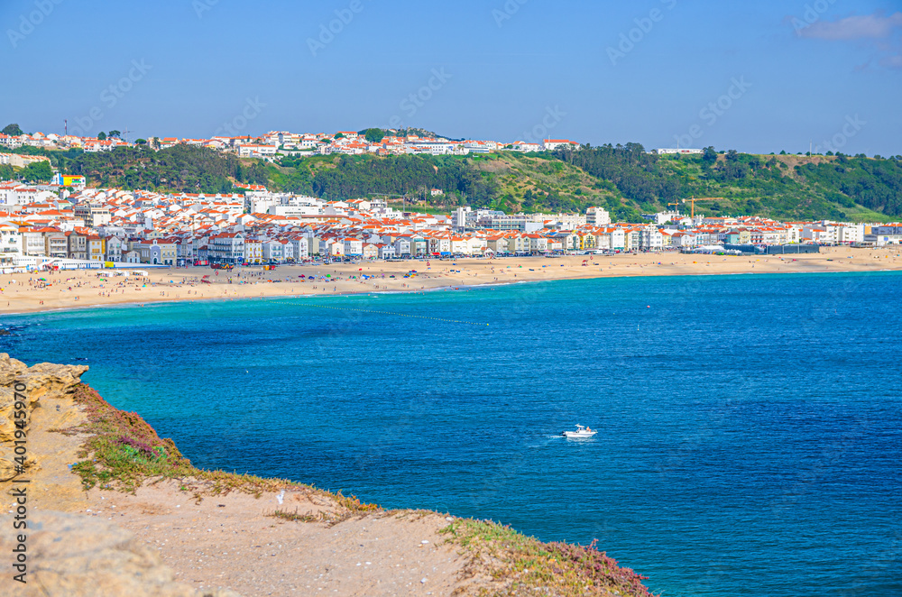 Aerial view of white yacht boat in azure turquoise water of Atlantic Ocean and sandy beach coastline Praia da Nazare town with small figures of people, Leiria District, Oeste region, Portugal