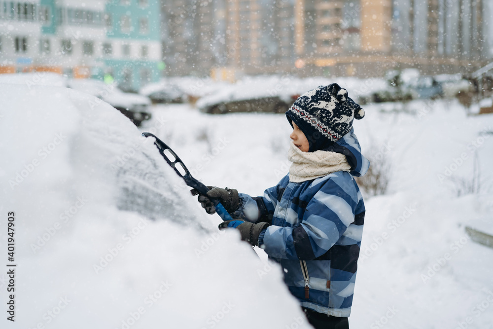 cute caucasian boy wearing scandinavian knit hat helping to brush snow from a car on winter day  near apartment building. Image with selective focus