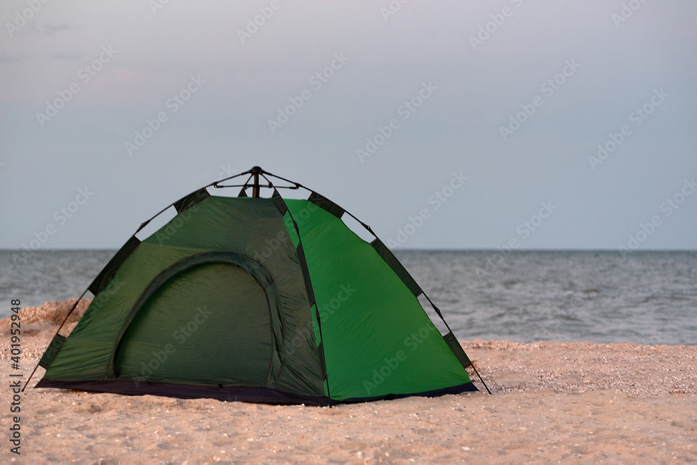 Green camping tent on sandy beach against sea background. Camping by the sea