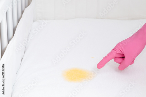 Hand in pink rubber protective glove pointing to yellow fresh urine stain on white mattress. Baby crib. Upholstered furniture cleanup. Closeup. photo