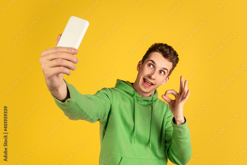 Selfie, vlog. Caucasian young man's portrait on yellow studio background. Beautiful male model in green outfit with cellphone. Concept of human emotions, facial expression, sales, ad, tech. Copyspace. Selfie, vlog. Caucasian young man's portrait on yellow studio background. Beautiful male model in green outfit with cellphone. Concept of human emotions, facial expression, sales, ad, tech. Copyspace.
