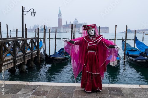Venice, Italy - February 18, 2020: An unidentified woman in a carnival costume in front of a group of gondolas and St Giorgio's Island,  attends at the Carnival of Venice. © Laia
