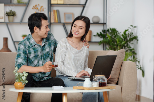 Smiling Asian man hugging girlfriend working on laptop at home and offering her a cup of tea