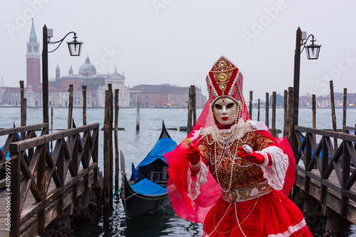 Venice, Italy - February 18, 2020: An unidentified woman in a carnival costume in front of a group of gondolas and St Giorgio's Island,  attends at the Carnival of Venice. © Laia