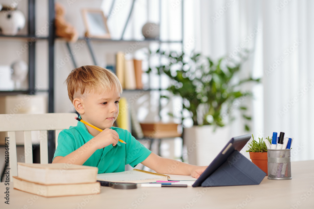 Smart little boy sitting at desk and watching education video on tablet computer or attending online class Smart little boy sitting at desk and watching education video on tablet computer or attending online class