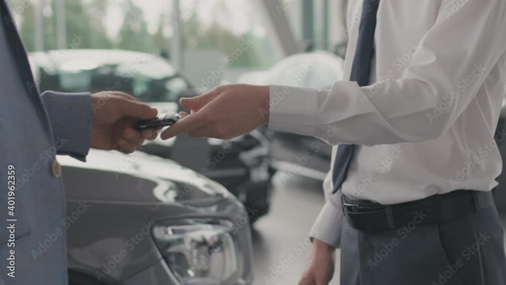 Mid-section close up of unrecognizable salesman in formal attire handing car keys to male customer and shaking hands in dealership after closing deal Mid-section close up of unrecognizable salesman in formal attire handing car keys to male customer and shaking hands in dealership after closing deal