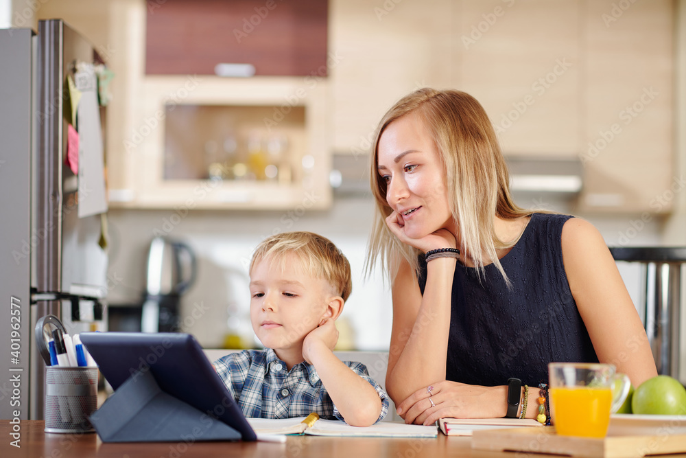 Mother and son watching webinar or online class on tablet computer when sitting at kitchen table at home Mother and son watching webinar or online class on tablet computer when sitting at kitchen table at home