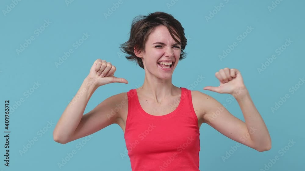 Surprised brunette young woman 20s in pink tank top posing isolated on blue background in studio. People lifestyle concept. Pointing index fingers on herself showing thumb up like gesture blinking Surprised brunette young woman 20s in pink tank top posing isolated on blue background in studio. People lifestyle concept. Pointing index fingers on herself showing thumb up like gesture blinking