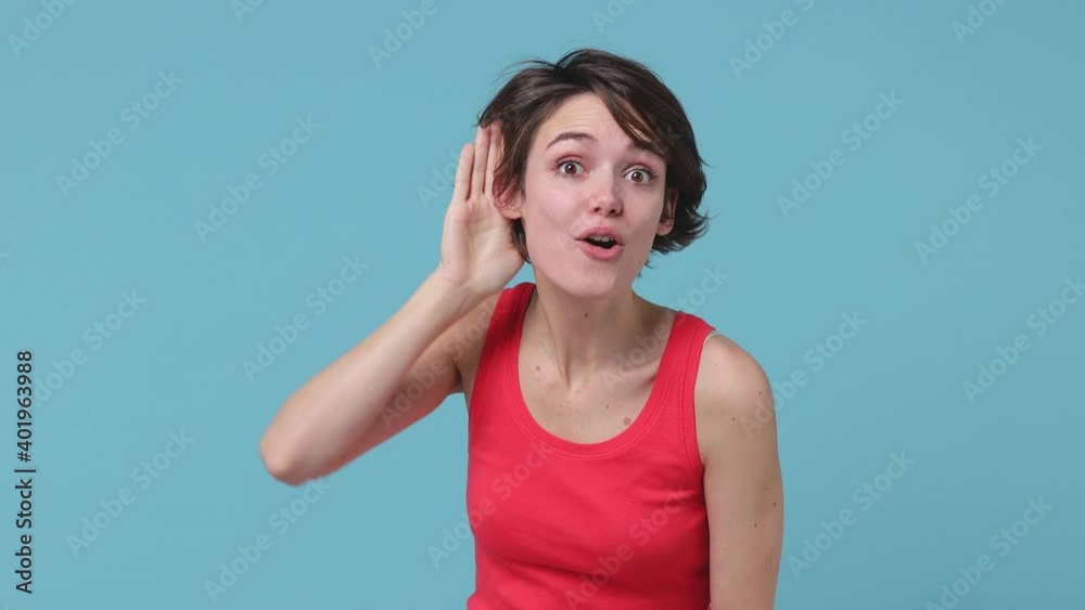 Curious shocked brunette young woman 20s years old in pink tank top posing isolated on pastel blue background in studio. People emotions lifestyle concept. Try to hear you overhear listening intently Curious shocked brunette young woman 20s years old in pink tank top posing isolated on pastel blue background in studio. People emotions lifestyle concept. Try to hear you overhear listening intently