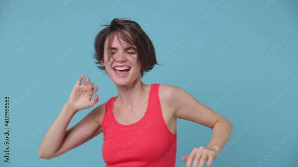 Cheerful funny excited young woman 20s in pink tank top posing isolated on blue background studio. People lifestyle concept. Dancing clenching fists waving hands pointing fingers showing victory sign