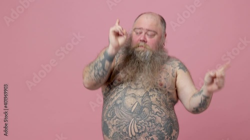 Cheerful fat pudge obese chubby overweight bearded man has tattooed naked bare big belly isolated on pink background. People lifestyle concept. Dancing clenching fists waving hands pointing fingers