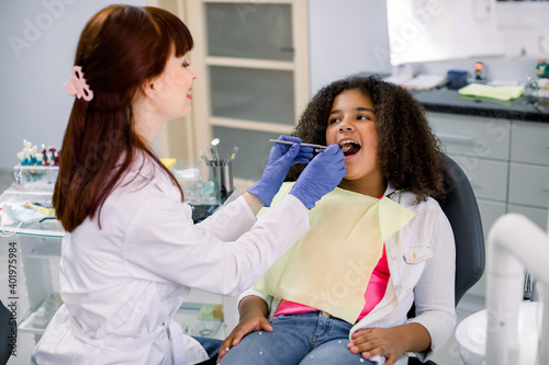 Pretty mixed raced teen girl in dentist chair, with mouth open, having dental examination. Side view of smiling female Caucasian dentist in gloves, holding dental tools