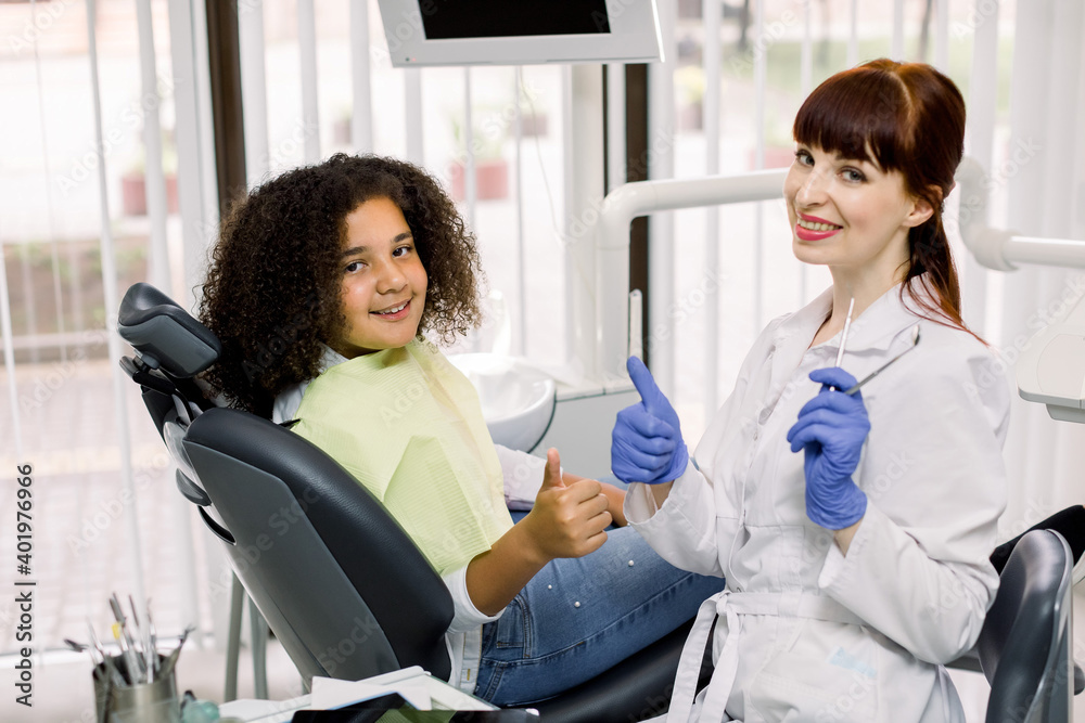Fototapeta premium Pediatric dentistry, teeth care and orthodontics. Young Caucasian woman dentist, showing thumbs up together with her little patient, mixed raced teen girl, sitting in dental chair in modern clinic
