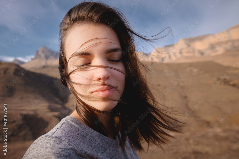 Fototapeta premium Portrait of an attractive Caucasian young woman in a checkered red shirt against the backdrop of mountains with eyes closed from pleasure. Bliss and pleasure in the mountains. The wind blows your hair