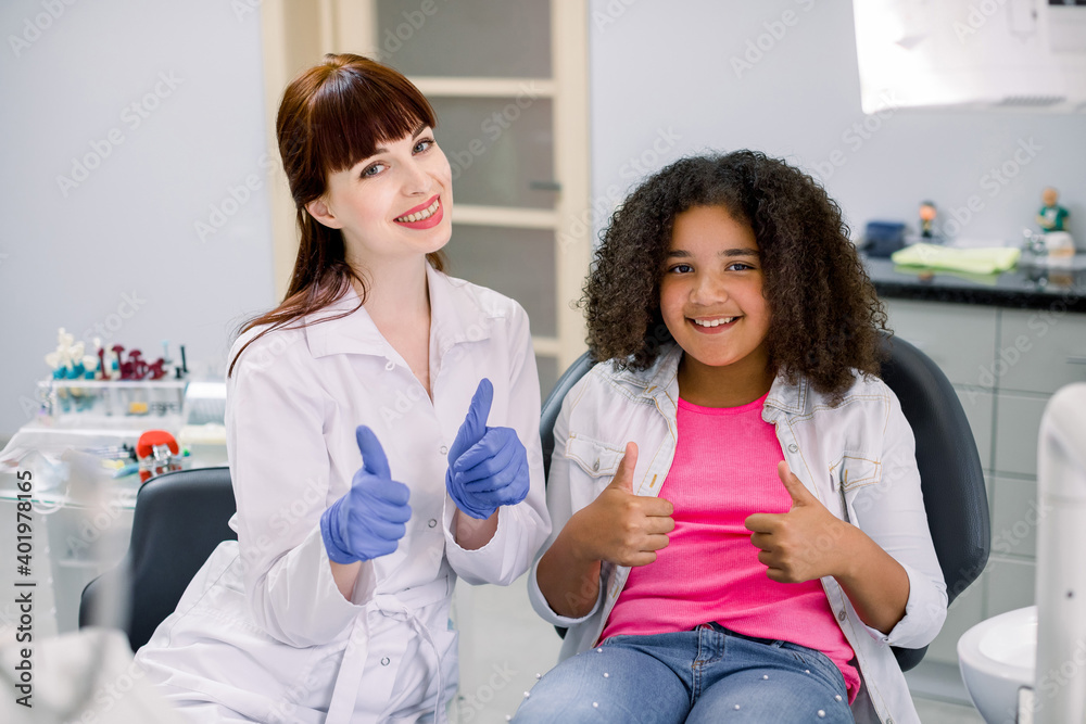 Excited happy mixed raced curly school girl, sitting on the chair at the dental office, together with her female smiling dentist, showing thumbs up and looking at camera Excited happy mixed raced curly school girl, sitting on the chair at the dental office, together with her female smiling dentist, showing thumbs up and looking at camera