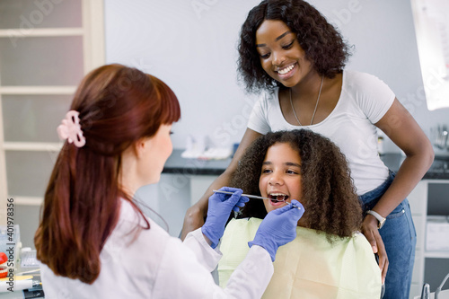 Dental treatment without fear. Female dentist in whilte uniform and gloves, examining teeth of little African American girl, while her mother supporting her behind, at modern pediatric dental clinic