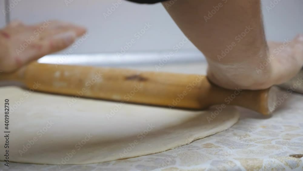 Young hands of chef rolling out pizza dough on a wooden surface at kitchen. Male arms of cook unrolling pastry using rolling pin on a cuisine table at restaurant. Food cooking concept. Close up