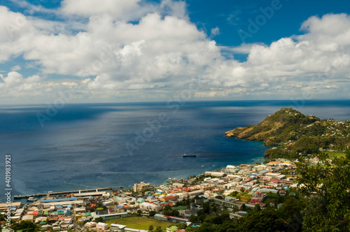 Fototapeta Naklejka Na Ścianę i Meble -  Houses of town in tropical bay. Coast, port, stadium and blue sea water with sky and white clouds. Aerial view of Kingstown, Saint Vincent. Caribbean lifestyle themes