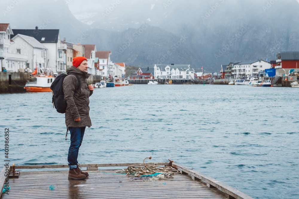 Traveler man with backpack standing on wooden pier on the background of lake and mountain houses. Space for your text message or promotional content. Travel lifestyle concept