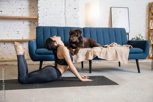 Calm. Young woman working out at home during lockdown, doing yoga exercises with the dog. Beautiful woman stretching, practicing. Wellness, wellbeing, healthcare, mental health, lifestyle concept.