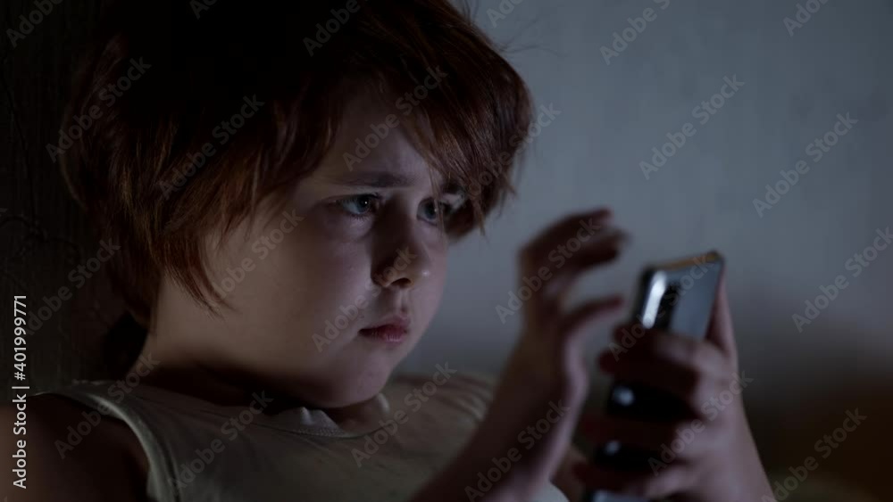 Portrait of a small girl with short hair and plump cheeks, in a t-shirt lying at home. She holds in her hands and uses a smartphone for games. Portrait of a small girl with short hair and plump cheeks, in a t-shirt lying at home. She holds in her hands and uses a smartphone for games.