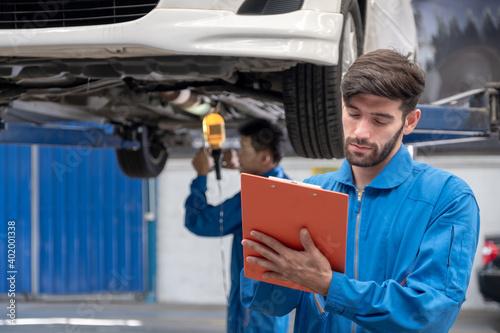 Professional Caucasian mechanic worker holding clipboard and checking list car fix items in front of car lifting with blurred mechanic on background.