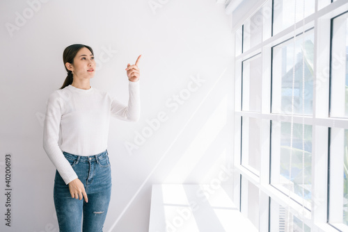 Good looking Asian female wearing a white sweater and jeans point at the wall near the window with natural light.