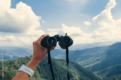 Hands holding binoculars on mountains forest nature background, looking through binoculars, travel, search and search concept.