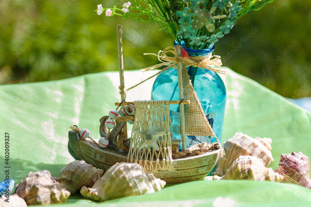 Sea thematic decorative elements for birthday party or event. Wooden toy ship, seashells, vase with flowers on green fabric background outdoors.