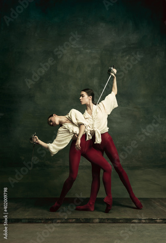 Passioned. Two young female ballet dancers like duelists with swords on dark green background. Caucasian models dancing together. Ballet and contemporary choreography concept. Creative art photo. © master1305