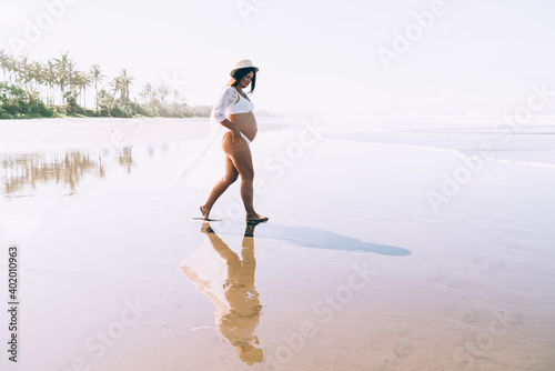 Trendy young pregnant woman walking along sandy seashore on sunny day