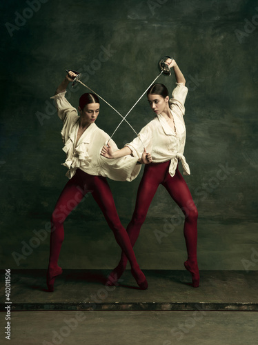 Contrast. Two young female ballet dancers like duelists with swords on dark green background. Caucasian models dancing together. Ballet and contemporary choreography concept. Creative art photo. © master1305