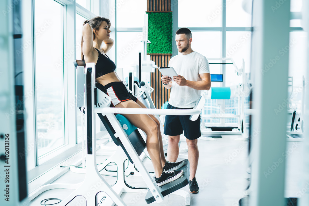 Instructor with tablet near sportswoman exercising on modern gym equipment