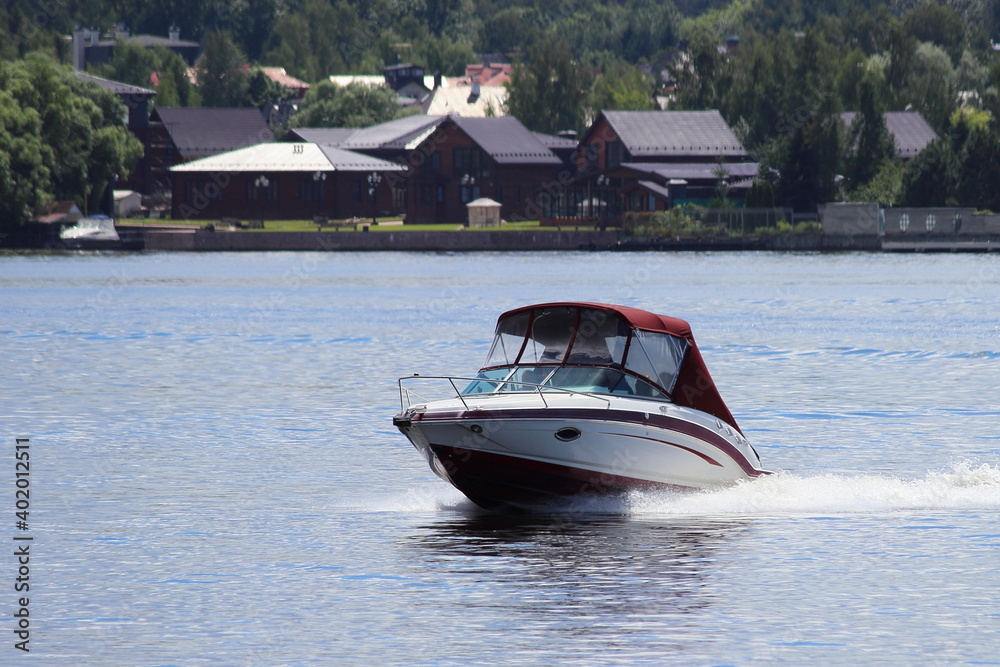 Fast gliding white motor boat front side view on cottage houses and green trees on far shore background in Moscow area at Sunny summer day, countryside active boating tourism recreation on river water