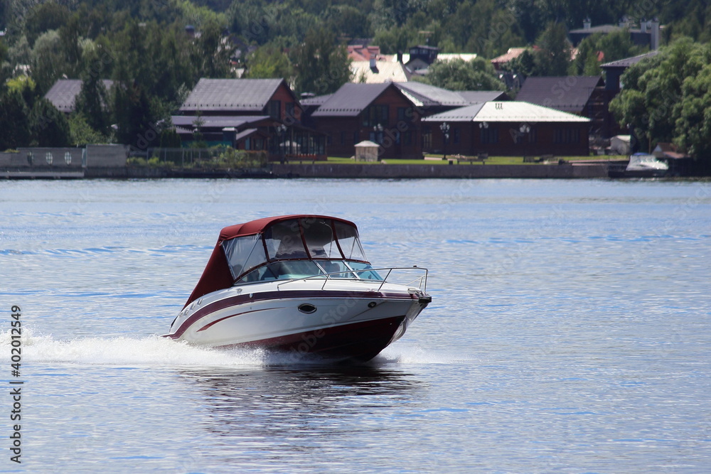 Fast floating white motor boat with closed red awning, front side view on green trees and cottages on far shore background at Sunny summer day, active recreation boating on lake water