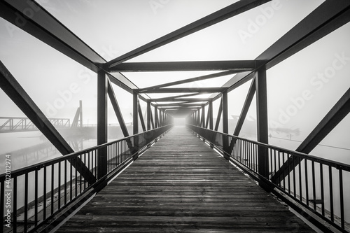 A grayscale shot of light at the end of a modern beautifully architectured bridge surrounded by fog photo