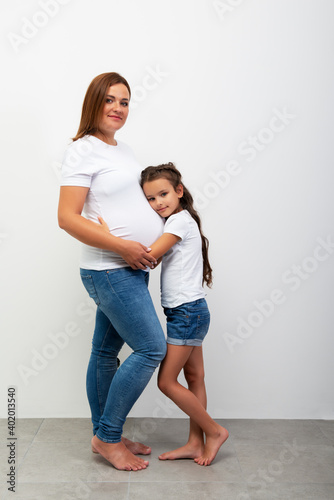 Pregnancy. Happy Family Expecting Baby. Beautiful smiling girl hugs her pregnant mother with big belly. Indoor portrait of mom and daughter. photo