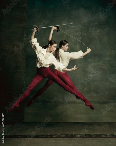 Fight. Two young female ballet dancers like duelists with swords on dark green background. Caucasian models dancing together. Ballet and contemporary choreography concept. Creative art photo. photo